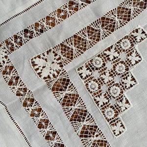 May include: Close-up of a white linen tablecloth with intricate lace and cutwork details. The design features geometric patterns and floral motifs, creating a delicate and elegant aesthetic. The tablecloth is ideal for special occasions.