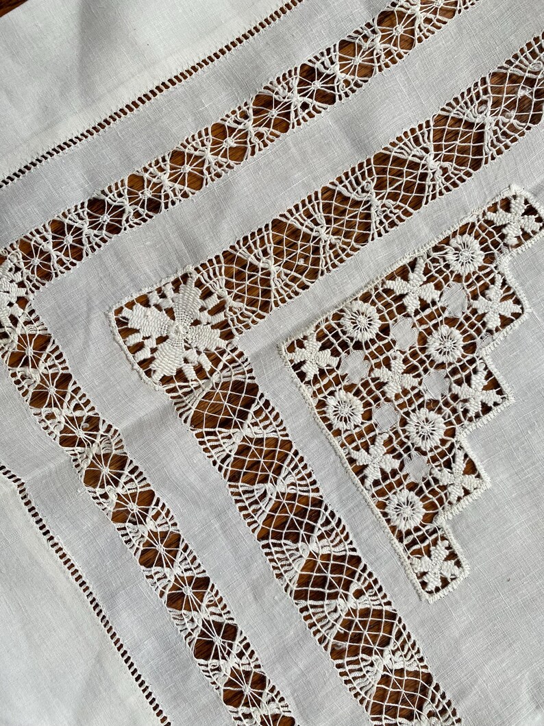 May include: Close-up of a white linen tablecloth with intricate lace and cutwork details. The design features geometric patterns and floral motifs, creating a delicate and elegant aesthetic. The tablecloth is ideal for special occasions.
