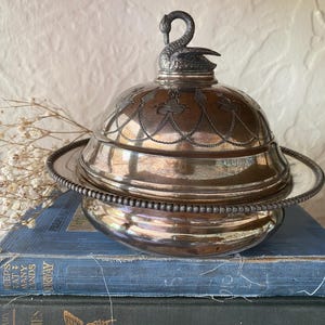 May include: A silver-toned metal butter dish with a domed lid featuring an engraved floral garland pattern and a sculpted swan finial handle. The base has a beaded rim edge, and the entire piece has a polished, reflective metallic finish.