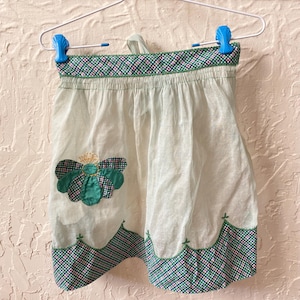 May include: A vintage-style, light green apron with a floral applique and a checkered pattern trim in green, pink, and white. The apron is hanging on a white hanger with blue clips, showcasing a retro kitchen accessory.