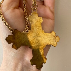 May include: A gold-toned cross pendant necklace with a detailed design. The back of the pendant is stamped with "LAMIA STERLING".