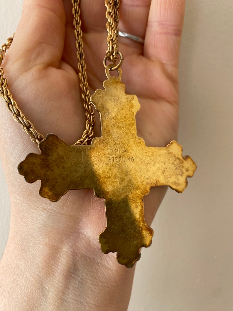 May include: A gold-toned cross pendant necklace with a detailed design. The back of the pendant is stamped with "LAMIA STERLING".