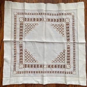 May include: A square, off-white linen tablecloth with intricate lace detailing. The border features a repeating geometric pattern with floral accents. The corners showcase a stepped design with delicate openwork.