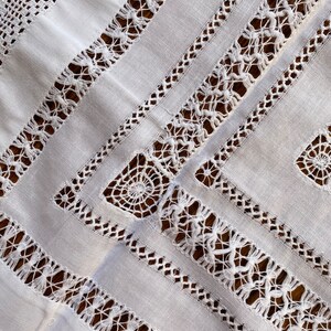 May include: White linen tablecloth with intricate crocheted lace trim. The lace features a geometric pattern with a repeating motif of small circles and squares.