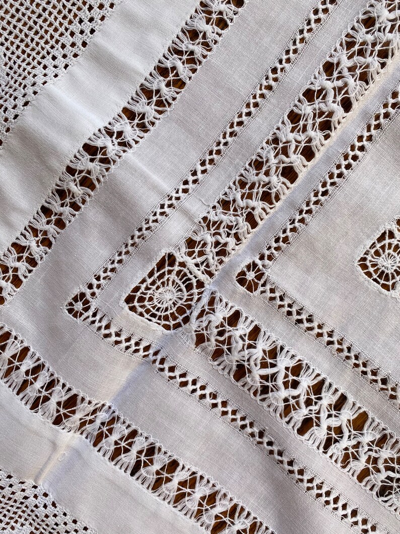 May include: White linen tablecloth with intricate crocheted lace trim. The lace features a geometric pattern with a repeating motif of small circles and squares.