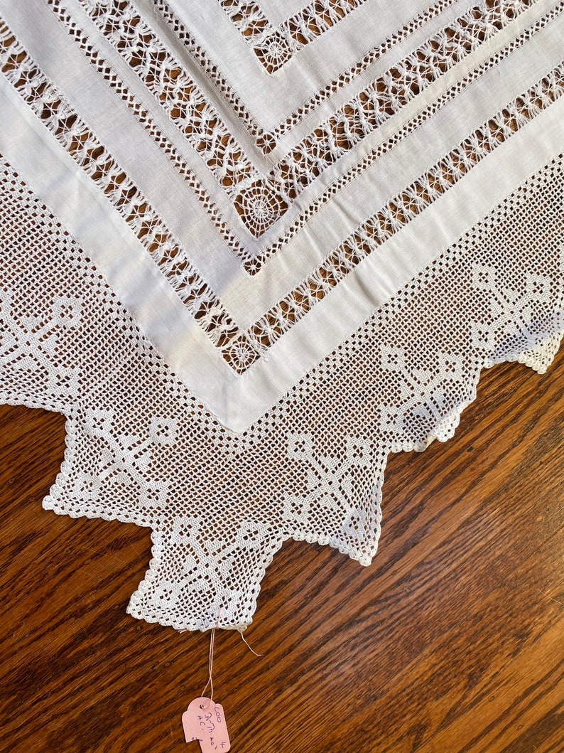 May include: White crocheted tablecloth with a square design in the centre and a scalloped edge. The tablecloth is on a wooden surface. The tag says "Crochet, 42" x 32", &pound;25.00"