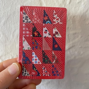 Puede incluir: Una baraja de cartas envuelta en plástico transparente. Los dorsos rojos de las cartas presentan un diseño de patchwork de triángulos con patrones florales y de lunares. Las cartas están sujetas en una mano.