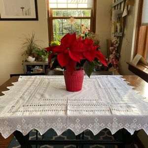 May include: A white crocheted tablecloth with a delicate lace pattern covers a wooden table. A red potted poinsettia plant sits in the centre of the table.
