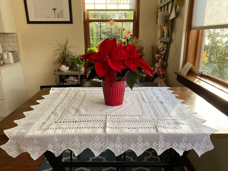 May include: A white crocheted tablecloth with a delicate lace pattern covers a wooden table. A red potted poinsettia plant sits in the centre of the table.