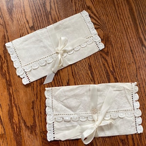 May include: Two rectangular off-white fabric envelopes featuring scalloped edges with eyelet embroidery details. Each envelope is secured with a matching satin ribbon tied in a bow at the center, with one ribbon featuring a small circular button detail.