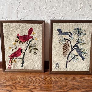 May include: Two framed punch needle wall art pieces featuring textured cream backgrounds. The left piece shows two red cardinals on a branch with yellow flowers. The right piece depicts two black and white birds on a pine branch with a pinecone. Both have the initials AP 81.