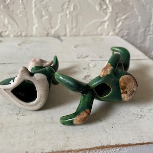 Op de afbeelding: Een keramieken beeldje van een zittende elf met groene kleding en een wit gezicht. De elf heeft een gat in zijn lichaam en ligt op zijn zij. Het beeldje heeft een verweerde, vintage uitstraling met blootliggende keramiekgebieden.