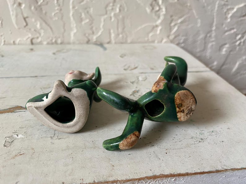 Op de afbeelding: Een keramieken beeldje van een zittende elf met groene kleding en een wit gezicht. De elf heeft een gat in zijn lichaam en ligt op zijn zij. Het beeldje heeft een verweerde, vintage uitstraling met blootliggende keramiekgebieden.