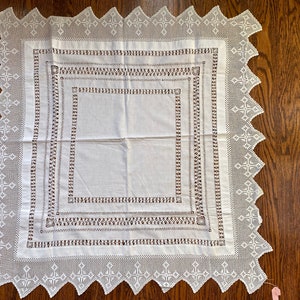 May include: White linen tablecloth with a delicate crocheted border and intricate embroidered details. The tablecloth features a square design with multiple layers of embroidery.