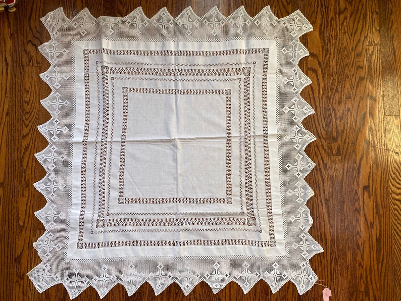 May include: White linen tablecloth with a delicate crocheted border and intricate embroidered details. The tablecloth features a square design with multiple layers of embroidery.