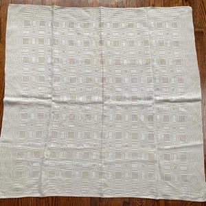 Op de afbeelding: Een rechthoekig, gebroken wit textiel met een herhalend geometrisch patroon van vierkanten en rechthoeken. De stof heeft een subtiele textuur en een licht, neutraal kleurenpalet. Het textiel is waarschijnlijk een tafelkleed of een decoratieve stof.