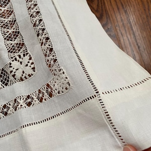 May include: Close-up of a white linen tablecloth with intricate lace and cutwork detailing. The edges are finished with a delicate, stitched border. The tablecloth is folded, revealing the texture of the fabric.