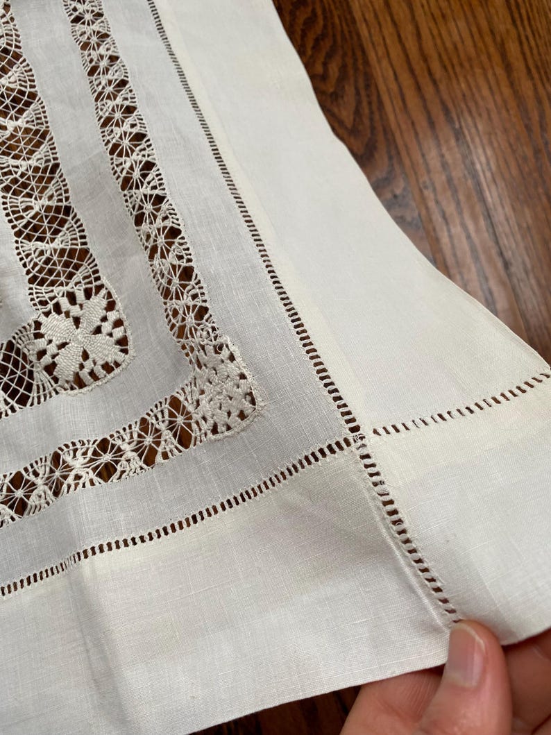 May include: Close-up of a white linen tablecloth with intricate lace and cutwork detailing. The edges are finished with a delicate, stitched border. The tablecloth is folded, revealing the texture of the fabric.