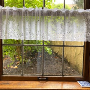 May include: A white lace window valance with a floral design hangs above a window. The valance is scalloped along the bottom edge. The window has a wooden frame and a view of a garden.