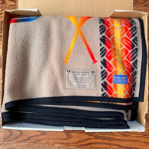 May include: A tan wool blanket with a black border, featuring a graphic of crossed orange paddles and a patterned stripe in red, orange, and yellow. A rectangular label reads STOMISH BLANKET 1946-1996, and a blue Beaver State brand tag is attached.