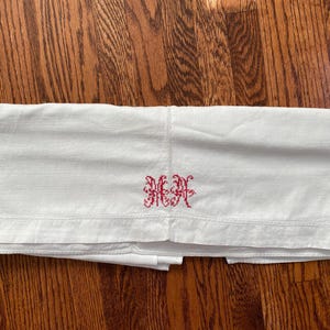May include: A white rectangular textile piece featuring a red embroidered monogram in the center. The fabric has a smooth texture with a visible hemmed border along the bottom edge. The red stitching forms two ornate, intertwined capital letters.