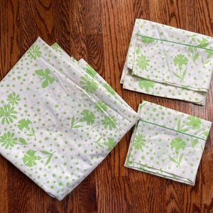 May include: White bed sheets and pillowcases with a light green floral pattern. The sheets are folded and arranged on a wooden surface. The floral design includes various flower shapes and small dots.