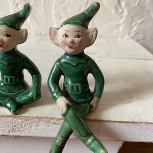 Op de afbeelding: Twee keramische elfjes. De elfen dragen groene outfits met bijpassende puntmutsen. Ze hebben een lichte huid, roze wangen en rode lippen. E&eacute;n elf zit met gekruiste benen en de andere in een vergelijkbare pose.
