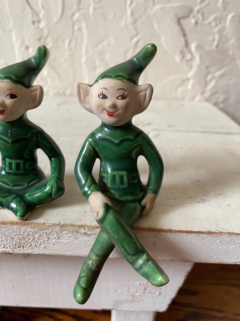 Op de afbeelding: Twee keramische elfjes. De elfen dragen groene outfits met bijpassende puntmutsen. Ze hebben een lichte huid, roze wangen en rode lippen. E&eacute;n elf zit met gekruiste benen en de andere in een vergelijkbare pose.