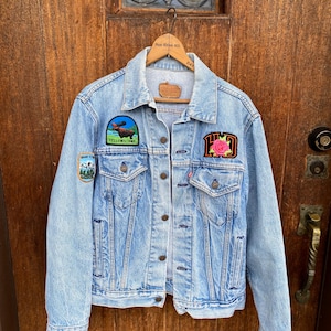 May include: Light blue denim jacket with patches. Patches include a rooster, a rose, and a moose. The jacket has a classic collar, button-up front, and two chest pockets. The jacket is hanging on a wooden hanger.