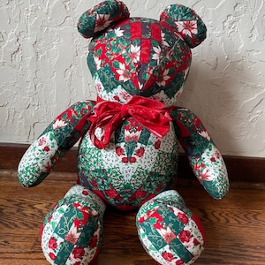 May include: A handmade teddy bear with a festive Christmas theme. The bear is crafted from fabric with a patchwork design featuring red, green, and white floral patterns. It has a red bow and is sitting on a wooden surface.