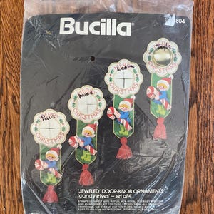Pode incluir: Um kit de ornamentos para maçaneta Bucilla Jeweled com quatro elfos de doces. O conjunto inclui feltro estampado, fio de bordar, fio de algodão e acabamentos decorativos. A embalagem mostra quatro ornamentos finalizados com o texto Merry Christmas.