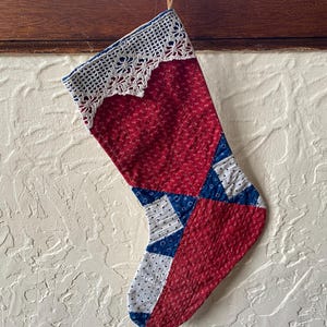 May include: A red, white, and blue patchwork Christmas stocking with a white crocheted trim. The stocking is quilted and has a blue and white geometric pattern.