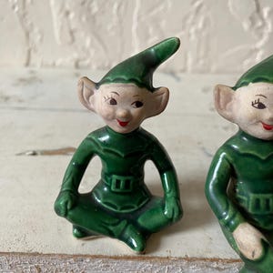Op de afbeelding: Twee keramische elfjes. De elfen dragen groene outfits en hoeden, met puntige oren en lachende gezichten. De beeldjes zitten met gekruiste benen. De achtergrond is een lichtgekleurd, getextureerd oppervlak.