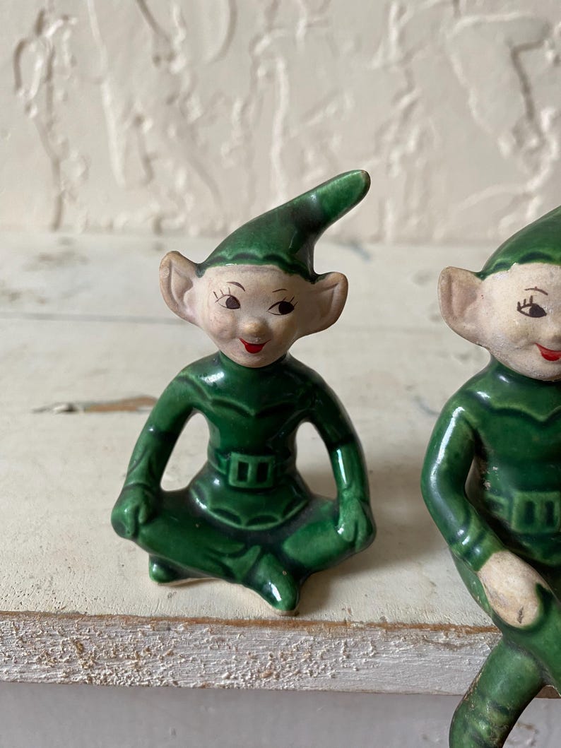Op de afbeelding: Twee keramische elfjes. De elfen dragen groene outfits en hoeden, met puntige oren en lachende gezichten. De beeldjes zitten met gekruiste benen. De achtergrond is een lichtgekleurd, getextureerd oppervlak.