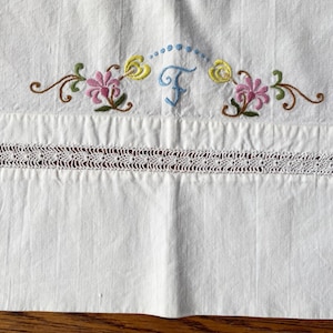 May include: White cotton pillowcase with embroidered flowers and the letter "F" in blue. The pillowcase has a lace trim.