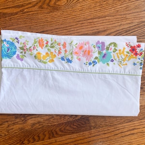 May include: A white pillowcase featuring a horizontal floral border pattern along the top edge. The print includes various flowers in shades of blue, orange, yellow, pink, purple, and red, accented by green foliage and a thin green horizontal line.