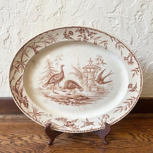 May include: An oval ceramic platter featuring a brown transferware pattern on a cream background. The central design depicts two turkeys standing near a small structure and a flying bird, surrounded by a border of floral and botanical motifs.