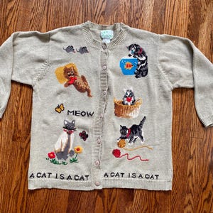 May include: A beige long-sleeve button-down cardigan featuring various cat-themed embroidered designs, including cats playing with yarn, a fishbowl, and mice. The text MEOW and A CAT IS A CAT is embroidered on the front of the knit garment.