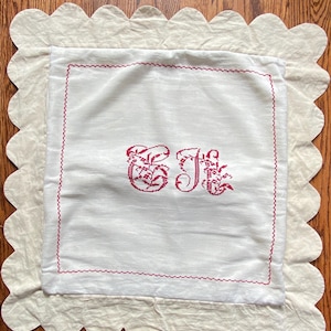 May include: A square white fabric cover featuring a scalloped border and a red cross-stitched monogram of the letters E and J in the center. A red stitched square frame surrounds the monogram, providing a decorative accent to the textile piece.