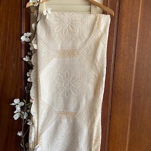 Pode incluir: Um tecido retangular de cor branco-sujo com um padrão repetitivo de medalhões florais bordados e painéis de renda geométrica. O tecido tem uma borda de renda festonada num dos lados e está pendurado num cabide de madeira.