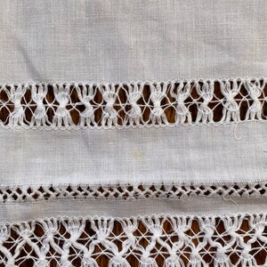 May include: White linen fabric with two rows of delicate white lace trim. The lace is a simple pattern of small knots and loops.