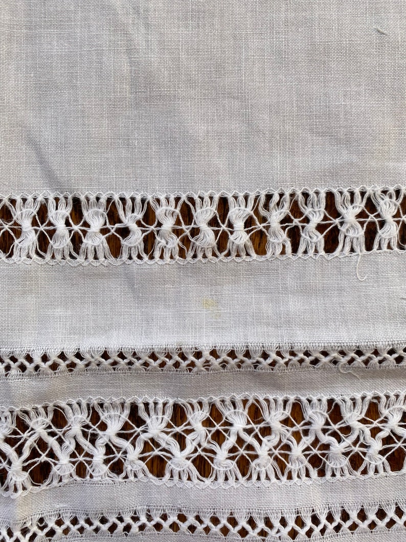 May include: White linen fabric with two rows of delicate white lace trim. The lace is a simple pattern of small knots and loops.