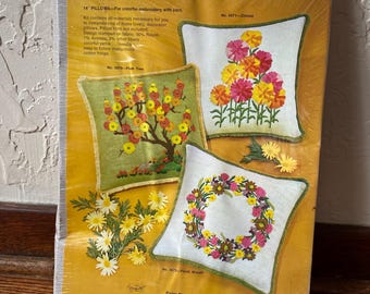 May include: A vintage embroidery kit for making 14 inch pillows. The kit includes materials for three different designs: a floral wreath, a tree with pink flowers, and a bouquet of pink and yellow flowers. The kit is from Fixler Bros., Inc. and is made in the USA.