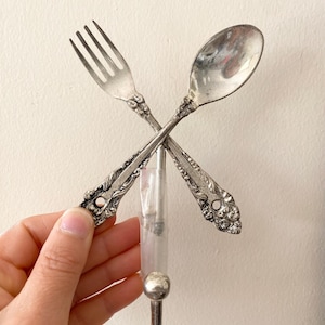 May include: A silver-toned wall hook featuring a decorative fork and spoon crossed at the handles. The metal utensils have an ornate, embossed floral pattern on the stems and include a single downward-facing hook at the base for hanging items.