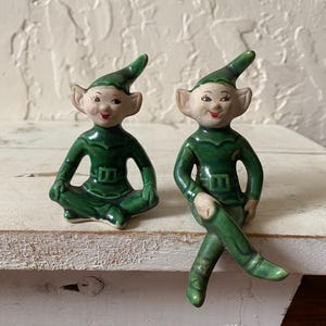 May include: Two ceramic elf figurines. One is seated cross-legged, the other has one leg crossed. Both are wearing green outfits with pointed hats and have rosy cheeks and red lips. The figurines are on a white surface.