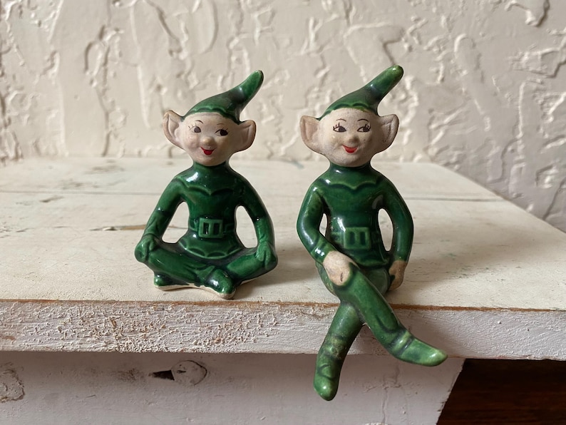 Op de afbeelding: Twee keramische elfenbeeldjes. E&eacute;n zit met gekruiste benen, de ander heeft &eacute;&eacute;n been gekruist. Beide dragen groene outfits met puntmutsen en hebben roze wangen en rode lippen. De beeldjes staan op een wit oppervlak.