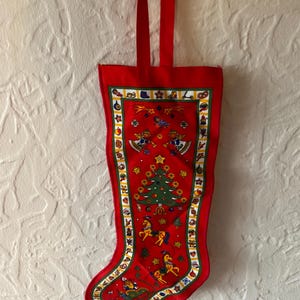 Può includere: Calza di Natale rossa con un design festivo. La calza presenta un albero di Natale, stelle e slitte trainate da cavalli. Un bordo verde e bianco incornicia lo sfondo rosso. La calza ha un anello rosso per appenderla.