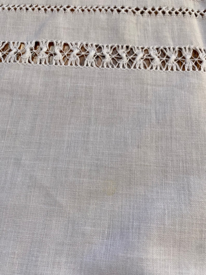 May include: A close-up of a white linen fabric with a delicate lace trim. The fabric has a subtle texture and a slightly off-white hue.