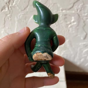 Op de afbeelding: Een kleine, groene keramische figuur van een kabouter of elf, van achteren gezien. De figuur heeft een puntmuts, grote oren en staat in een loopstand. Het keramiek heeft een glanzende afwerking met wat slijtage.