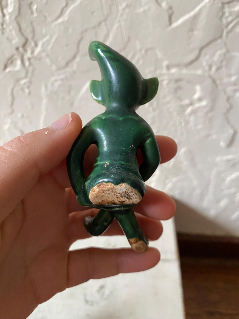 Op de afbeelding: Een kleine, groene keramische figuur van een kabouter of elf, van achteren gezien. De figuur heeft een puntmuts, grote oren en staat in een loopstand. Het keramiek heeft een glanzende afwerking met wat slijtage.